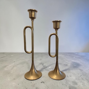 May include: Two brass candle holders shaped like trumpets, each featuring a tall, slender stem, a flared base, and a cup at the top for holding a candle. The metal has a smooth, polished finish with a warm, golden tone and a distinct loop design.