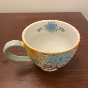 May include: A ceramic mug with a floral design. The mug features a light blue handle and a yellow exterior with a floral pattern in shades of blue, pink, and orange. The rim is outlined in orange.