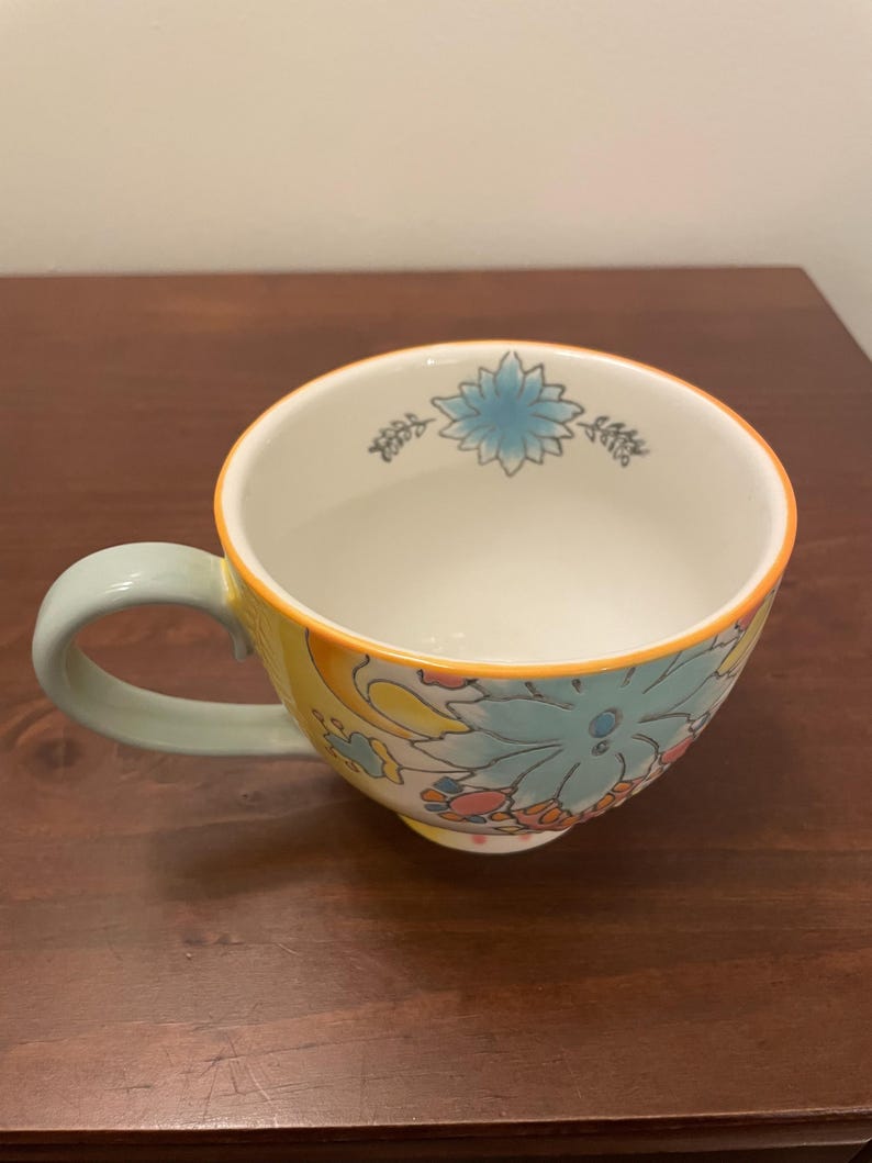 May include: A ceramic mug with a floral design. The mug features a light blue handle and a yellow exterior with a floral pattern in shades of blue, pink, and orange. The rim is outlined in orange.