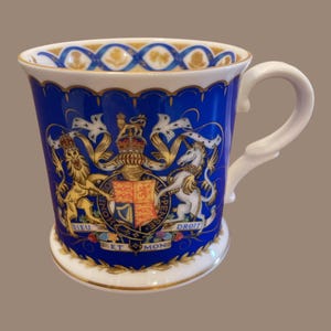 Może przedstawiać: Niebiesko-biały ceramiczny kubek z dekoracyjnym herbem i złotymi zdobieniami. Kubek posiada biały uchwyt i szczegółowy herb z koroną, lwem i jednorożcem. Obręcz i podstawa mają złoto-niebieski wzór.