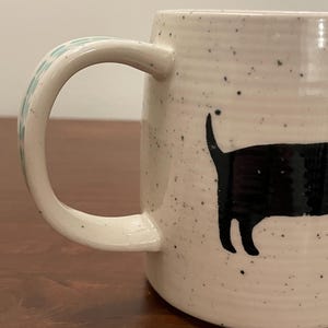 May include: Cream-colored ceramic mug with a black cat silhouette design. The mug has a speckled finish and a curved handle with a light blue floral pattern. The cat is depicted with its tail up, adding a playful touch to the design.