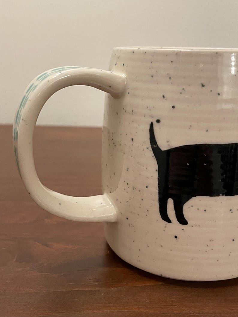 May include: Cream-colored ceramic mug with a black cat silhouette design. The mug has a speckled finish and a curved handle with a light blue floral pattern. The cat is depicted with its tail up, adding a playful touch to the design.