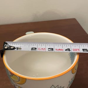 May include: White ceramic bowl with floral patterns in blue, yellow, and green, and an orange rim. The bowl's diameter is approximately 4 inches. A measuring tape is placed across the bowl.