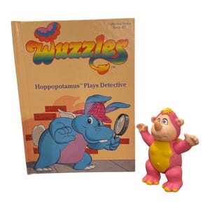 Könnte beinhalten: Ein Vintage-Kinderbuch mit dem Titel "Wuzzles Hoppopotamus Plays Detective" mit einem blauen Nilpferd-Charakter auf dem Cover und einer rosa-gelben Wuzzles-Spielfigur. Das Buch gehört zur Collector-Serie.
