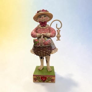 Pode incluir: Uma estatueta extravagante de um gato vestido com um vestido floral rosa e um chapéu de palha, segurando uma cesta cheia de flores. O gato está sobre uma base verde com um design de coração, e um coelhinho decorativo pendurado.