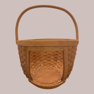 May include: A light brown woven basket with a curved handle. The basket has a rounded base and a rectangular opening. The basket is made of woven wood strips and has a natural finish.