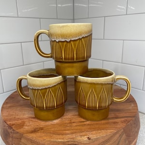 Könnte beinhalten: Drei braune Keramikbecher mit geometrischem Muster. Die Becher haben eine hellbraune Glasur oben und eine dunkelbraune Glasur unten. Die Becher sind übereinander gestapelt.