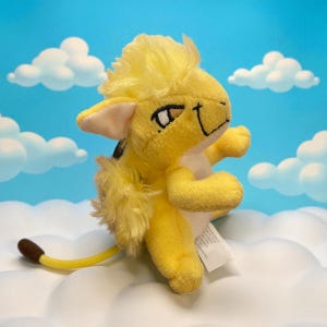 May include: A plush yellow lion cub toy with a fluffy mane, tail, and chest. The toy has black eyes and a smiling expression. The background is a blue sky with white clouds.