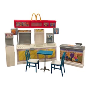 Pode incluir: Um conjunto de brinquedos de restaurante de fast-food em miniatura com um letreiro vermelho e amarelo, um balcão e uma mesa com duas cadeiras. O conjunto inclui um dispensador de bebidas, uma caixa registradora e expositores de comida. O letreiro tem o logotipo dos arcos dourados.