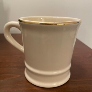 May include: A white ceramic mug with a gold rim. The mug has a rounded shape and a handle.