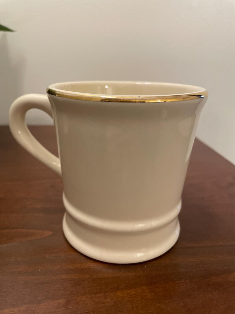 May include: A white ceramic mug with a gold rim. The mug has a rounded shape and a handle.