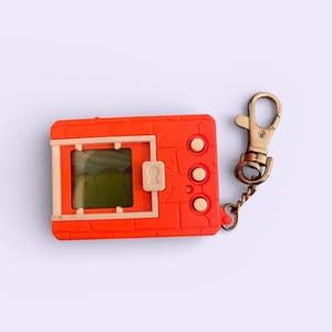 May include: An orange handheld digital pet game with a small screen displaying a green landscape. It has three white buttons and a key ring attachment. The device is a retro toy.