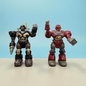 May include: Two toy robots. One is black, brown, and gold, with a drill arm and antenna. The other is red and grey, with a cannon arm. Both have articulated limbs and are posed against a light blue background.