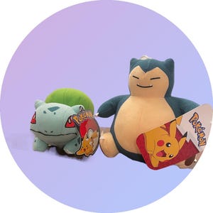 May include: Two plush Pokemon toys, a green and blue Bulbasaur and a brown and blue Snorlax. Both toys are sitting on a light purple background. The toys have a Pokemon tag attached to them.