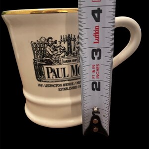 May include: White ceramic mug with a gold rim and a black and white image of a barber shop with the text "Paul Moore" and "1031 Lexington Avenue/New York, NY Established 19..."
