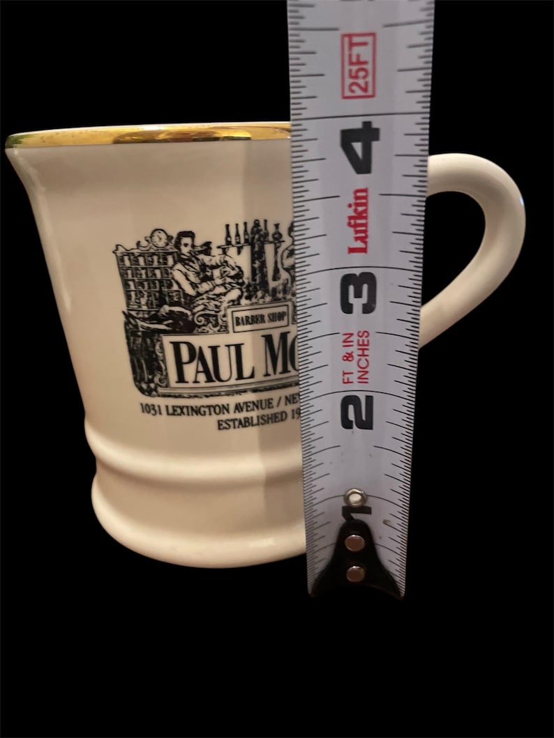 May include: White ceramic mug with a gold rim and a black and white image of a barber shop with the text "Paul Moore" and "1031 Lexington Avenue/New York, NY Established 19..."