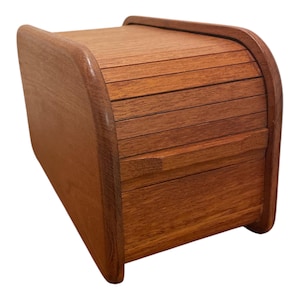 May include: A medium-brown wooden storage box featuring a curved top and a sliding tambour door mechanism. The box has a smooth, polished finish with visible wood grain patterns, rounded side panels, and a rectangular base structure.