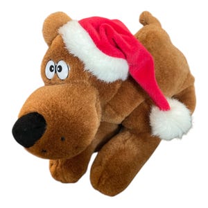 May include: A brown plush dog toy with large cartoon eyes and a black nose. The dog is wearing a red Santa hat with white trim and a white pom-pom. The toy is designed to look like a cartoon character.