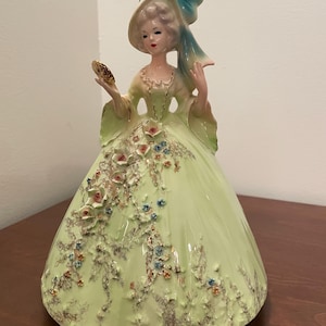 May include: A ceramic figurine of a woman in a long, light green dress with floral accents. She wears a hat with a blue bow and holds a small mirror. The figurine has a vintage aesthetic.