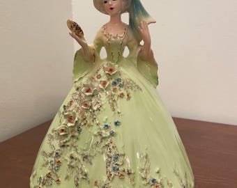May include: A ceramic figurine of a woman in a long, light green dress with floral accents. She wears a hat with a blue bow and holds a small mirror. The figurine has a vintage aesthetic.