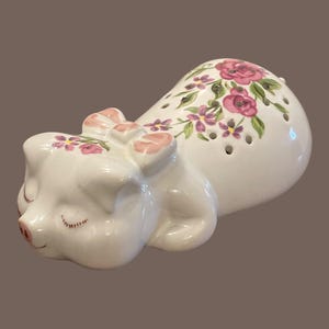 May include: A white ceramic pig-shaped salt and pepper shaker set. The pig is lying down with its eyes closed, and a pink bow on its head. The shaker has a floral design with pink and purple flowers.