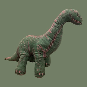 May include: A green plush dinosaur toy with pink accents. The long-necked dinosaur has a detailed pattern of pink spots and lines across its body. The toy is standing on a solid green background.