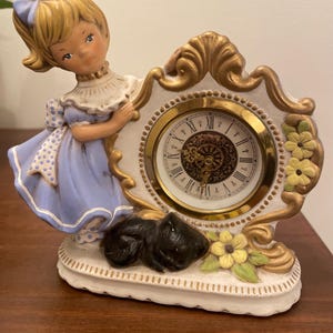 May include: A vintage ceramic clock featuring a girl in a blue dress, leaning against a white and gold clock face. The clock has Roman numerals and is adorned with yellow flowers and a black cat figurine. The clock measures approximately 6 inches in height.
