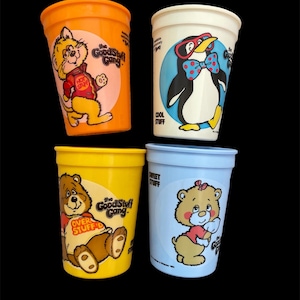 May include: Set of four colorful plastic cups featuring cartoon characters from the "Good Stuff Gang". Each cup has a different character: a cat, a penguin, a bear, and a bear with a bow. The cups are all decorated with the "Good Stuff Gang" logo.