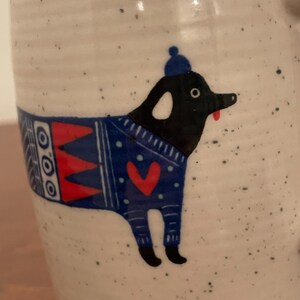 May include: A speckled ceramic mug featuring a whimsical illustration of a black dog wearing a blue sweater with a red heart and a matching blue hat. The dog has a red tongue sticking out. The mug has a handle and a white background.