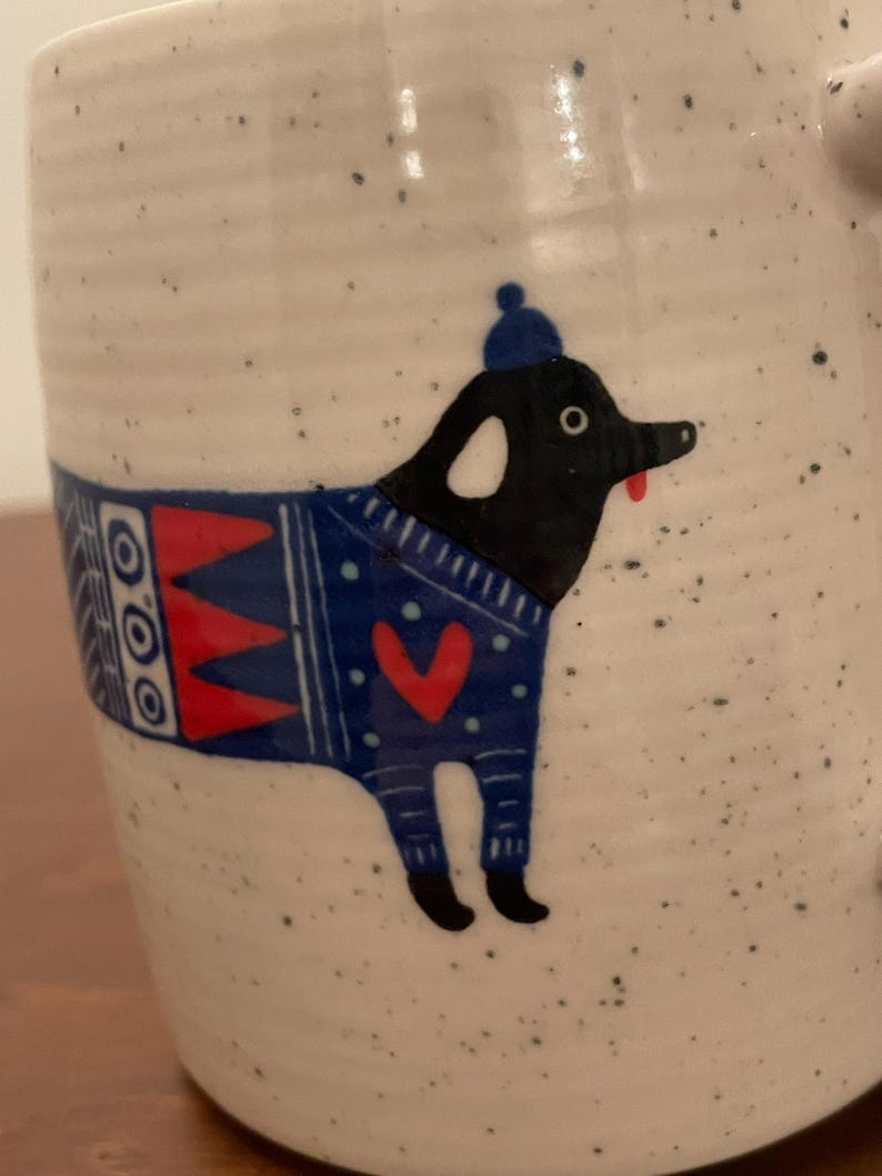May include: A speckled ceramic mug featuring a whimsical illustration of a black dog wearing a blue sweater with a red heart and a matching blue hat. The dog has a red tongue sticking out. The mug has a handle and a white background.
