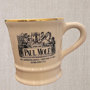 May include: A white ceramic mug with a gold rim and black lettering. The mug features a vintage illustration of a barbershop with the text "Paul Mole Barber Shop 1031 Lexington Avenue / New York, NY 10021 Established 1913."