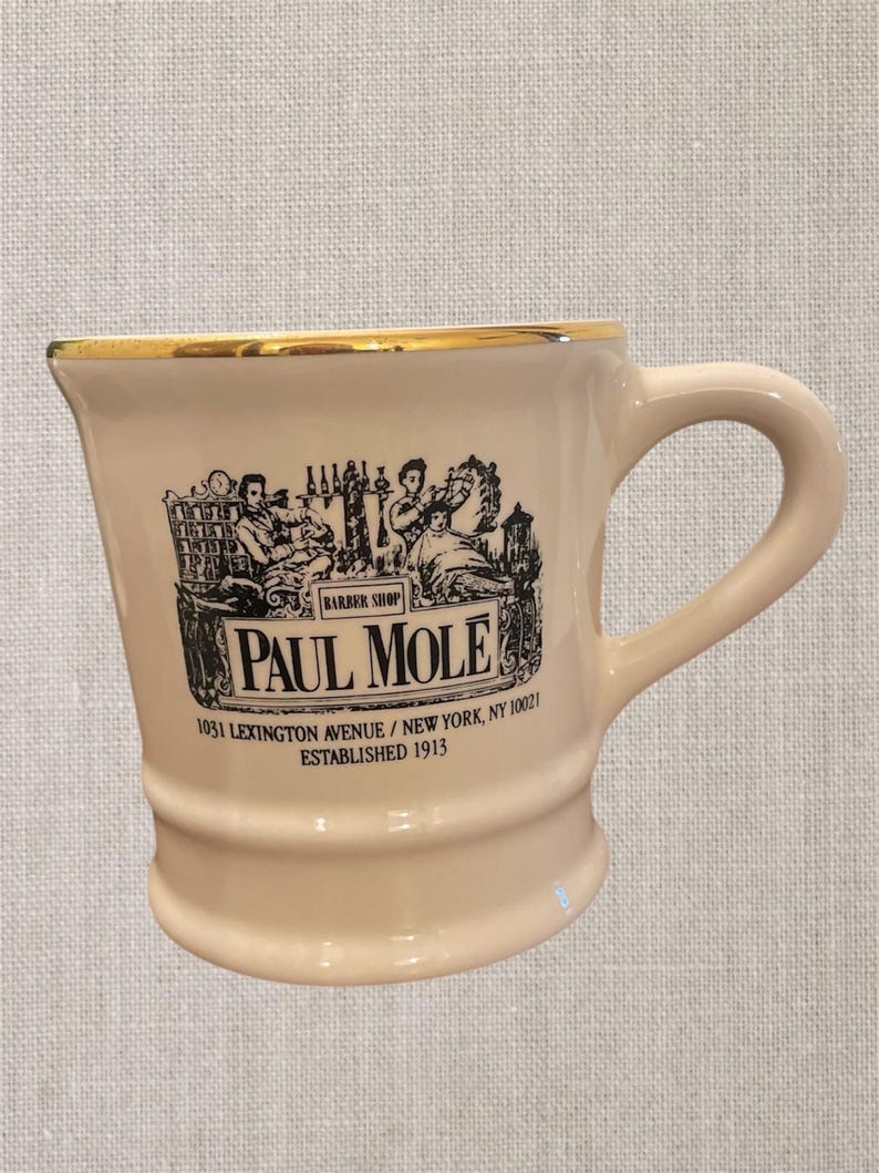 May include: A white ceramic mug with a gold rim and black lettering. The mug features a vintage illustration of a barbershop with the text "Paul Mole Barber Shop 1031 Lexington Avenue / New York, NY 10021 Established 1913."