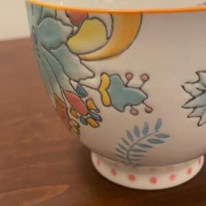 May include: White ceramic bowl with floral and leaf designs in blue, yellow, and pink. The rim is outlined in orange, and the base features pink polka dots. A decorative piece for serving or display.