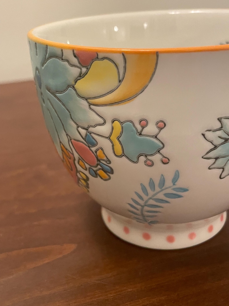 May include: White ceramic bowl with floral and leaf designs in blue, yellow, and pink. The rim is outlined in orange, and the base features pink polka dots. A decorative piece for serving or display.
