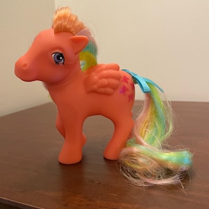 May include: A vintage orange toy pony with a rainbow mane and tail. The pony has a blue ribbon on its tail and pink star accents on its wings. The pony is standing on a wooden surface.