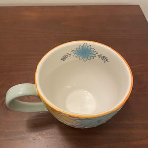 May include: A ceramic coffee mug with a light green handle and a floral design. The mug features a white interior, an orange rim, and a blue and yellow floral pattern on the exterior. The mug is sitting on a wooden surface.