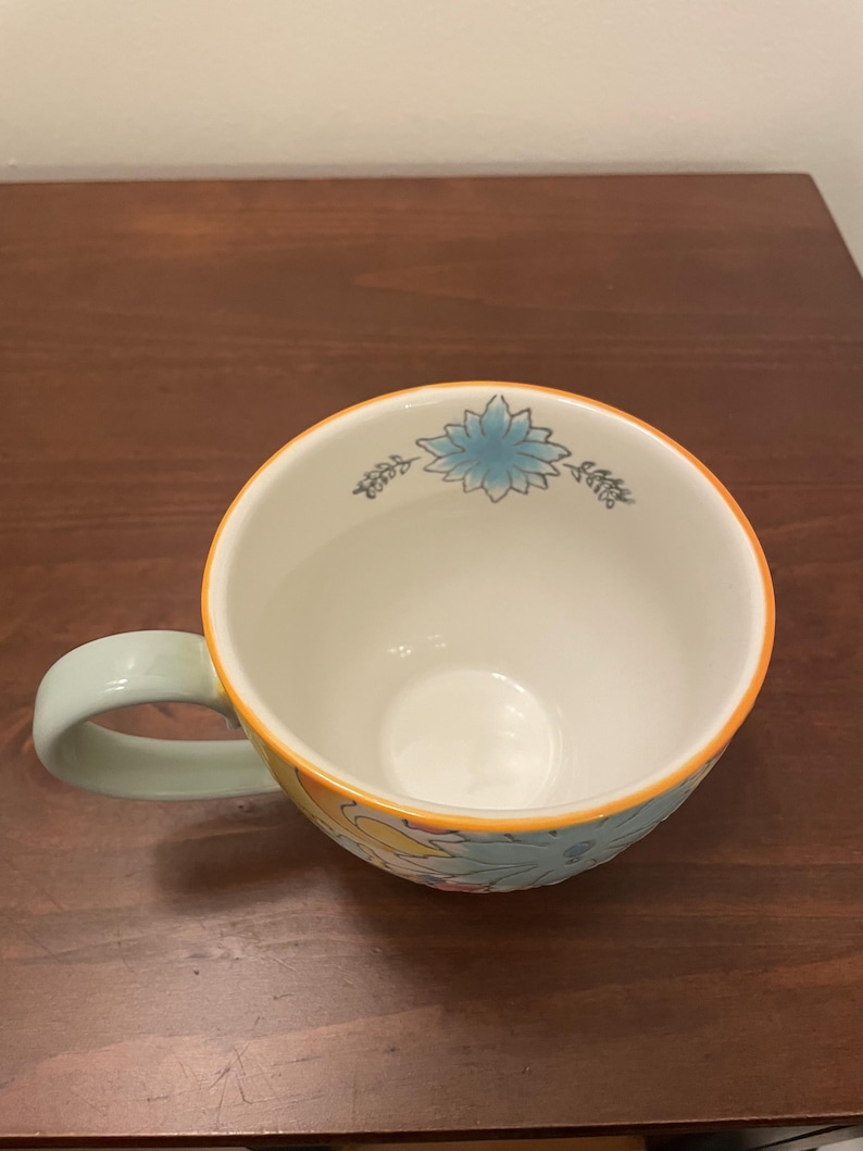 May include: A ceramic coffee mug with a light green handle and a floral design. The mug features a white interior, an orange rim, and a blue and yellow floral pattern on the exterior. The mug is sitting on a wooden surface.
