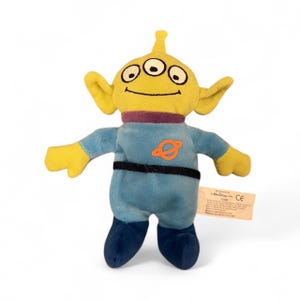 Könnte beinhalten: Ein Plüschtier-Alien aus dem Film Toy Story. Das Alien hat drei Augen, gelbe Haut und große Ohren. Es trägt einen blauen Overall mit einem orangefarbenen Planetenemblem und einem schwarzen Gürtel. Das Spielzeug hat einen lila Kragen und blaue Füße.