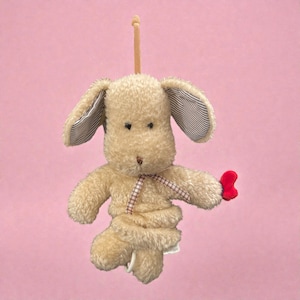 May include: A plush dog toy in cream with striped ears and a red bone. The toy has a red and white checkered ribbon around its neck and a long beige string for hanging. The background is a solid pink.