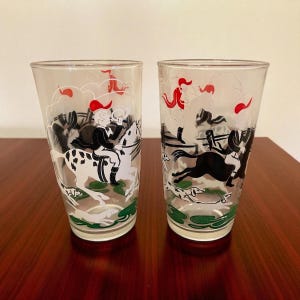 May include: Two clear drinking glasses with a hunting scene design. The glasses feature illustrations of riders on horseback, hounds, and red accents. The base of the glasses has a green border.