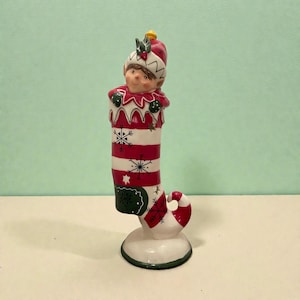May include: A ceramic Christmas elf figurine. The elf is wearing a red and white striped stocking with snowflake and star details. The elf has a red hat with holly leaves and a smiling face. The base is white with a green trim.
