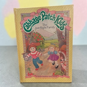 May include: A hardback book titled Cabbage Patch Kids The Just-Right Family, A Parker Brothers Story Book by Larry Callen with pictures by Judy Callen. The yellow cover features an illustration of two children running through a forest path.