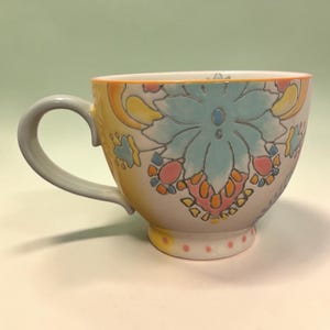 May include: A ceramic teacup with a floral design. The cup features a light blue floral pattern with pink, yellow, and orange accents. The handle is gray, and the rim is orange. The cup sits on a white surface.