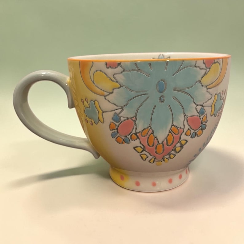 May include: A ceramic teacup with a floral design. The cup features a light blue floral pattern with pink, yellow, and orange accents. The handle is gray, and the rim is orange. The cup sits on a white surface.