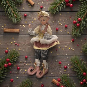 May include: A Christmas elf figurine with a cream-colored hat and outfit, accented with silver and pink details. The elf is posed on a wooden surface, surrounded by pine branches, red berries, cinnamon sticks, and gold confetti.