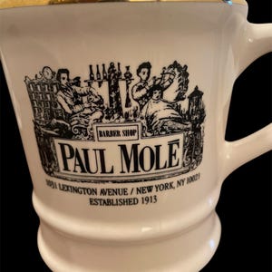 May include: White ceramic mug with a gold rim and black illustration of a barbershop scene. The mug has the text "BARBER SHOP", "PAUL MOLE", "1031 LEXINGTON AVENUE / NEW YORK, NY 10021", and "ESTABLISHED 1913" printed on it.