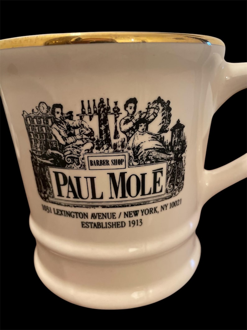 May include: White ceramic mug with a gold rim and black illustration of a barbershop scene. The mug has the text "BARBER SHOP", "PAUL MOLE", "1031 LEXINGTON AVENUE / NEW YORK, NY 10021", and "ESTABLISHED 1913" printed on it.