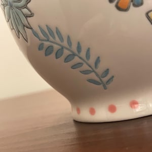 May include: Close-up of a white ceramic bowl with floral and leaf designs in blue, pink, and orange. The base features pink polka dots. The bowl is on a brown surface.