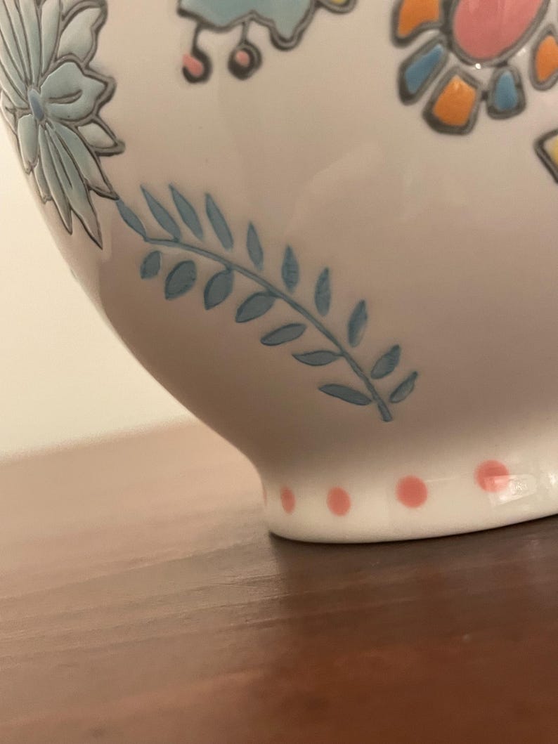 May include: Close-up of a white ceramic bowl with floral and leaf designs in blue, pink, and orange. The base features pink polka dots. The bowl is on a brown surface.