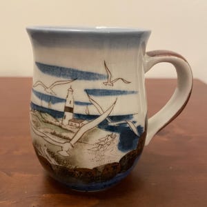 Pode incluir: Caneca de cerâmica com uma cena costeira com um farol, gaivotas e ondas do mar. A caneca tem uma borda e alça azuis, com um detalhe marrom na alça. A obra de arte é em tons de azul, marrom e branco.